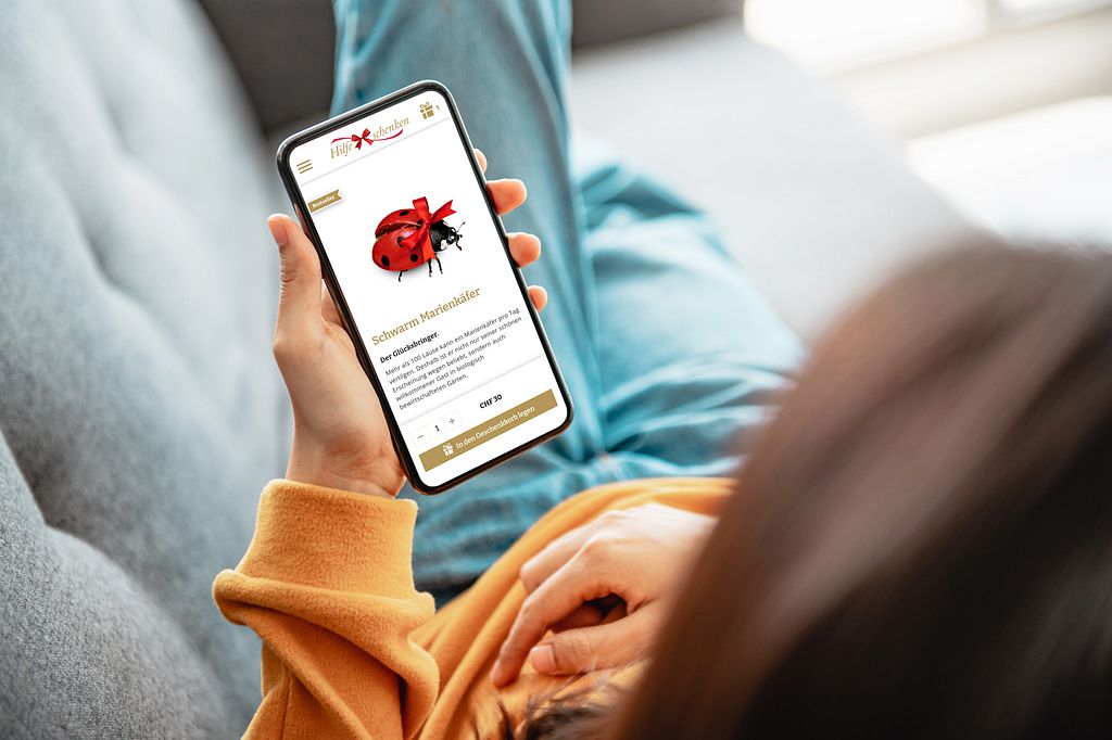Frau liegt auf dem Sofa und schaut sich den Webshop von «Hilfe schenken» auf dem Smartphone an